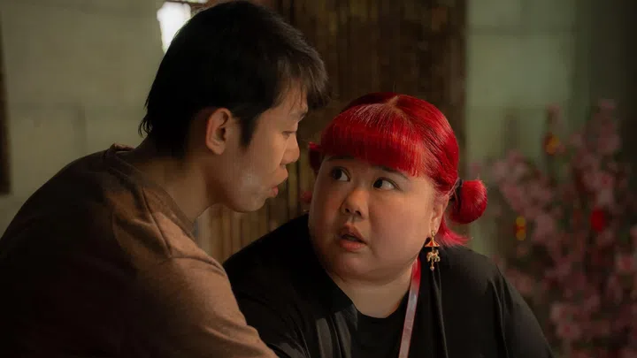 《百万红包 A Good Fortune》电影剧照，一名男子靠近女主角耳边低声说话，女主角染着红色双马尾发型，神情惊讶又带着疑惑地回望对方，背景可见室内新年装饰与粉色花卉布置，整体光线温暖，营造出紧张中带点温情的家庭氛围，呈现角色之间关键对话与情绪转折的瞬间。