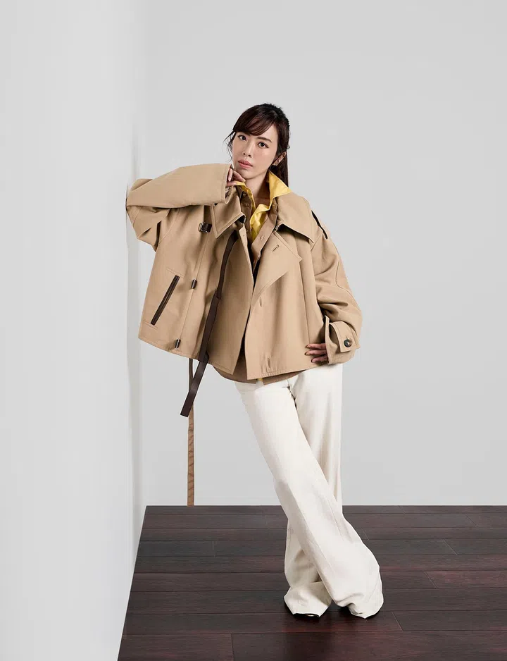 一位女性站在深色木地板上，背景为简洁的浅灰色墙面，整体画面干净利落。她将身体微微倾向墙面，一只手轻触下巴，另一只手放在腰间，姿态随性中带点自信与从容。她留着深棕色长发，绑成低马尾，妆容精致，眼神坚定地看向镜头。穿着卡其色宽松外套，内搭亮黄色衬衫，下身搭配米白色阔腿长裤与浅色高跟鞋，整体造型以中性色为主，搭配亮色点缀，展现出现代女性利落、优雅又具个性的时尚风格。