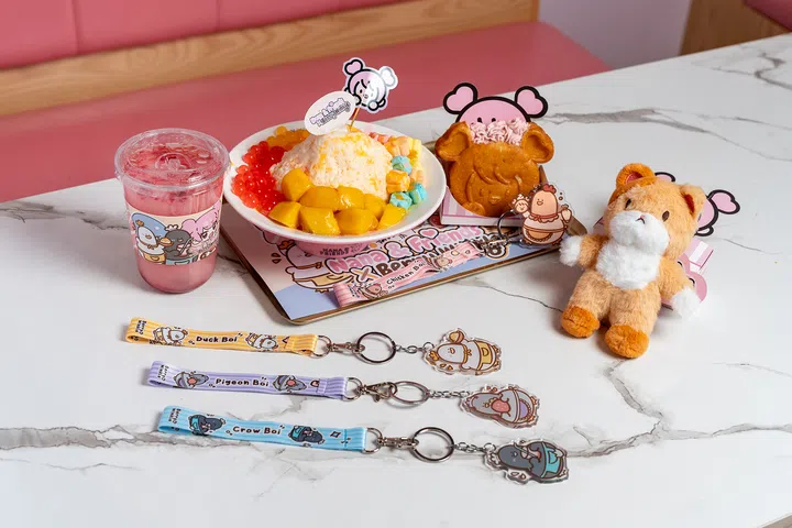 Nana & Friends与新加坡插画家Berry Winkle的IP Bird Bois联名产品展示。图中摆放在咖啡馆大理石桌上的餐点包括草莓饮品、芒果与彩色配料的刨冰（bingsu）、以及印有Nana & Friends角色图案的松饼甜点。周边产品有毛绒玩偶、钥匙扣和角色挂绳，分别印有Duck Boi、Pigeon Boi和Crow Boi卡通形象。