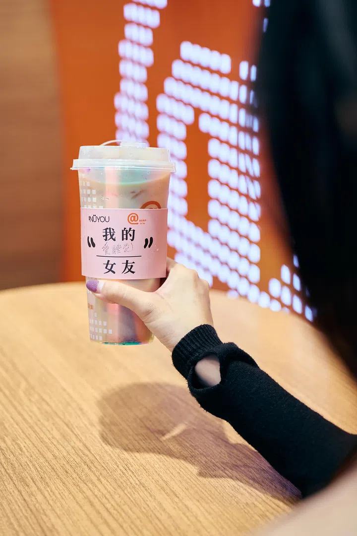 一名参加者手持《女友》与At Tea联名推出的“女友莓果伯爵”饮品。杯身套有粉色联名杯套，印有“我的女友”字样及《女友》和At Tea标志，背景为橙色主题布置。