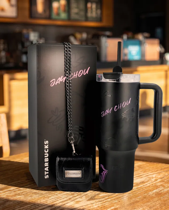 Jay Chou x Starbucks黑色手柄吸管杯与联名礼盒摆放在咖啡店木桌上。杯身印有粉紫色“JAY CHOU”字样，并饰有钢琴等音乐主题图案，顶部配透明杯盖和黑色吸管。礼盒前方放着带Starbucks金属铭牌的黑色迷你小包，深色链条垂挂于盒身，背景为虚化的咖啡店吧台。