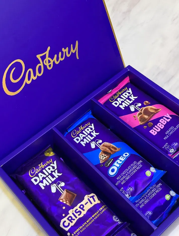 一个紫色 Cadbury 巧克力礼盒打开摆放在浅色大理石桌面上，盒盖内侧印有金色的“Cadbury”品牌标志。礼盒内整齐排列三款 Cadbury Dairy Milk 巧克力，包括紫色包装的 Crisp-It、蓝色包装的 Oreo，以及粉色包装的 Bubbly，每款巧克力分别放在独立的分隔格中，包装设计色彩鲜明，展示不同口味的牛奶巧克力产品。2026情人节礼物推荐：10款精致礼物让TA一秒心动