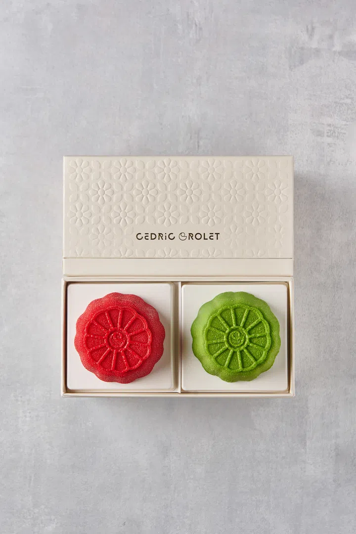 一只米白色礼盒，盒面压印花朵图案，并印有 “Cédric Grolet” 字样。礼盒内整齐放置两枚造型精致的月饼，分别为鲜红色与翠绿色，饼面呈花瓣放射状纹理，整体风格简洁雅致。Cedric Grolet Singapore，2025中秋月饼必买推荐。