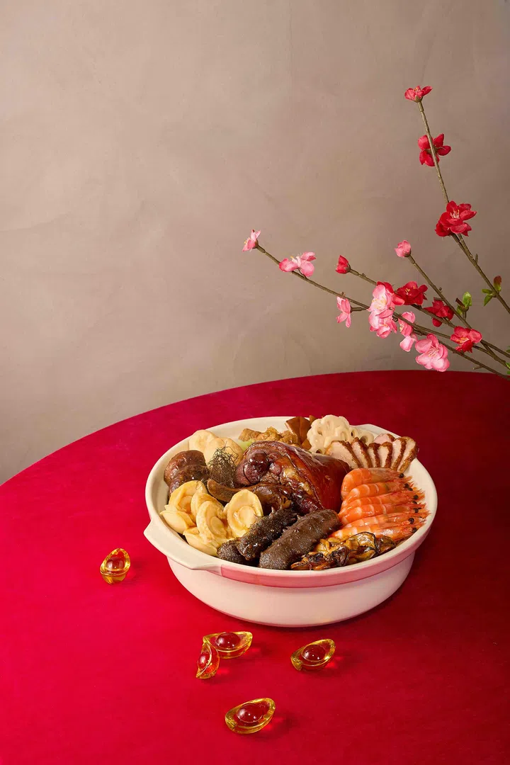 红色桌面上摆放一盆传统盆菜，白色砂锅中铺排整虾、鲍鱼、海参、花胶、猪脚与莲藕等多款海味与肉类，旁边点缀金元宝摆饰与梅花枝装饰，营造喜庆热闹的农历新年团圆饭氛围，新加坡盆菜推荐2026 农历新年必吃餐厅与价格整理。