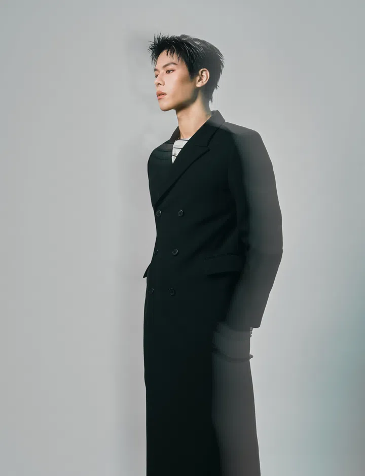 容启航Glenn Yong为《女友》拍摄时尚肖像，身穿黑色双排扣长外套，内搭黑白横条纹上衣，双手置于身后，侧身望向画面左方。他的黑色短发向上梳起，神情沉静，浅灰色背景与柔和光影衬托出侧脸轮廓，画面带有朦胧的重影效果。