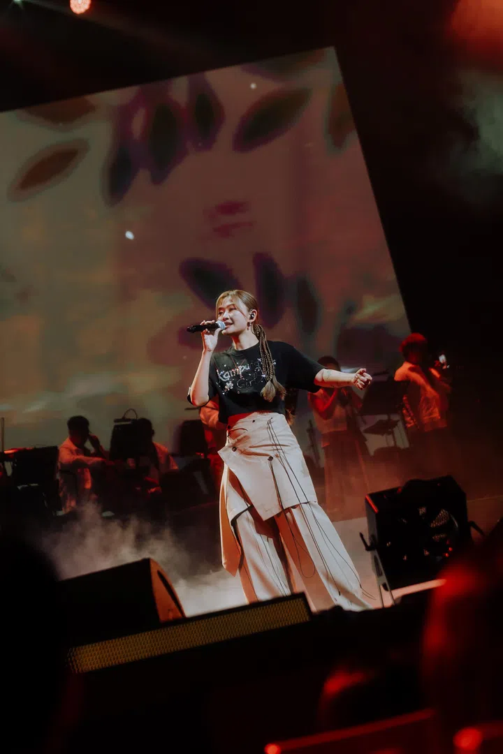 DIOR大颖在新加坡演唱会舞台上演唱，身穿黑色上衣搭配米色长裙造型，手持麦克风投入表演，肢体自然摆动与观众互动。舞台以暖红色灯光与烟雾营造氛围，后方大型屏幕呈现抽象人像视觉，与现场乐手同框，构成层次丰富的现场画面，展现她自由且富感染力的舞台表现。