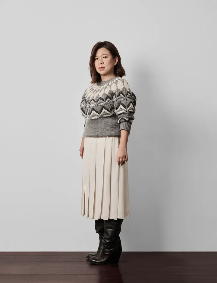 一位女性站在浅灰色背景前，穿着灰白色几何图案毛衣、浅色百褶中长裙和黑色长靴，身体微侧，正面对镜头。