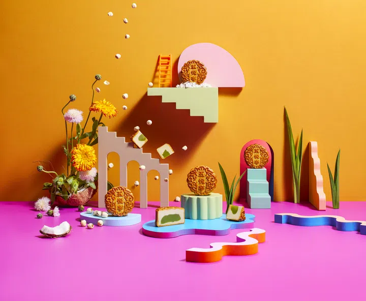 Andaz Singapore 的 Pandan Jade 月饼以艺术化布景拍摄：橙黄与亮粉背景下，几枚金黄烘焙月饼摆放在几何造型的彩色底座上，部分切开可见翠绿色夹心。画面点缀椰子、莲子与鲜花，构成趣味十足的超现实风格场景。Andaz Singapore，,2025中秋月饼必买推荐。