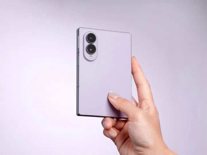一只手单手握着Samsung Galaxy Z Fold8 Lavender（薰衣草紫），展示折叠状态下的机身背面设计。手机采用雾面淡紫色背板，搭配双镜头相机模组及LED闪光灯，整体线条简洁俐落，镜头模组与机身同色设计，展现低调质感。使用者以拇指轻托机身，突显Galaxy Z Fold8轻巧纤薄、适合单手握持的特点。背景采用淡紫色渐层搭配柔和灯光，进一步衬托手机的精致外观与现代设计风格。