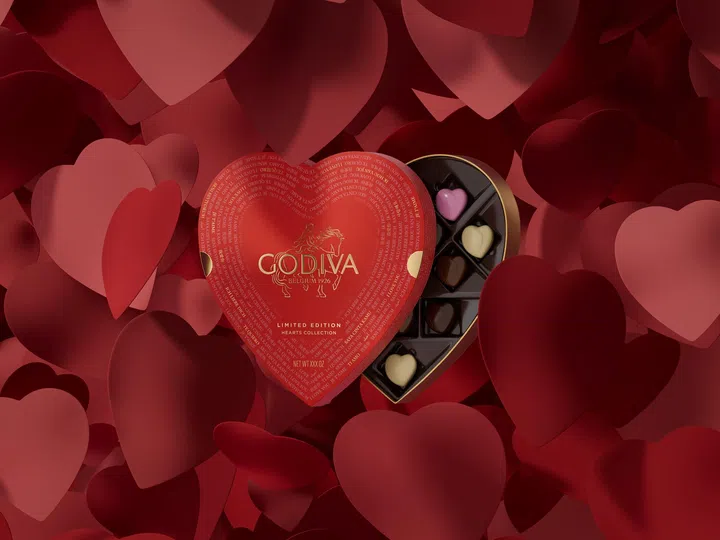 GODIVA 情人节限定心形巧克力礼盒置于红色爱心背景中央，整体画面充满浓厚的节日氛围。礼盒采用经典红色心形设计，盒盖印有金色 GODIVA 标志与骑马女神图案，下方标示“Limited Edition Hearts Collection”字样，展现品牌的精致与标志性风格。礼盒微微打开，可见内部整齐排列的心形巧克力，包括粉色、米色与深棕色不同口味，每颗巧克力均呈现光滑细腻的表面，造型精致。背景由不同层次与大小的红色爱心图案组成，营造出立体感与浪漫氛围，使礼盒成为视觉焦点，突显其作为经典情人节礼物的象征意义。2026情人节礼物推荐：10款精致礼物让TA一秒心动