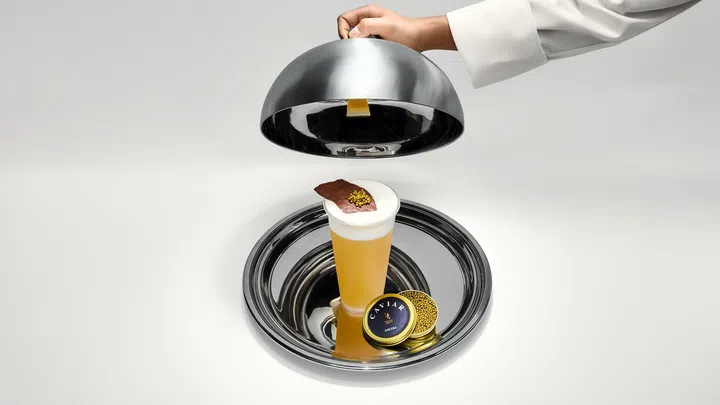 一杯呈金黄色的茶饮放在银色托盘中，表面覆盖奶泡和一片巧克力脆片，上方点缀少量鱼子酱，旁边摆放着金色鱼子酱罐，一只手正掀起银色圆顶餐盖，画面高级精致。