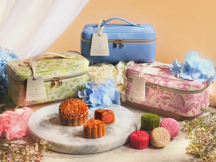 The Ritz-Carlton, Millenia Singapore 2026中秋Mid-Autumn Artistry Collection场景图，画面展示三款可重复使用的月饼vanity case礼盒，包括浅蓝色、绿色植物花纹及粉色花卉图案设计，均配有拉链、手柄及The Ritz-Carlton吊牌。前景陈列多款传统烘焙月饼与彩色冰皮月饼，其中一颗切开的烘焙月饼可见咸蛋黄内馅；周围以蓝色、白色及粉色花卉装饰，整体呈现柔和雅致的中秋送礼氛围。