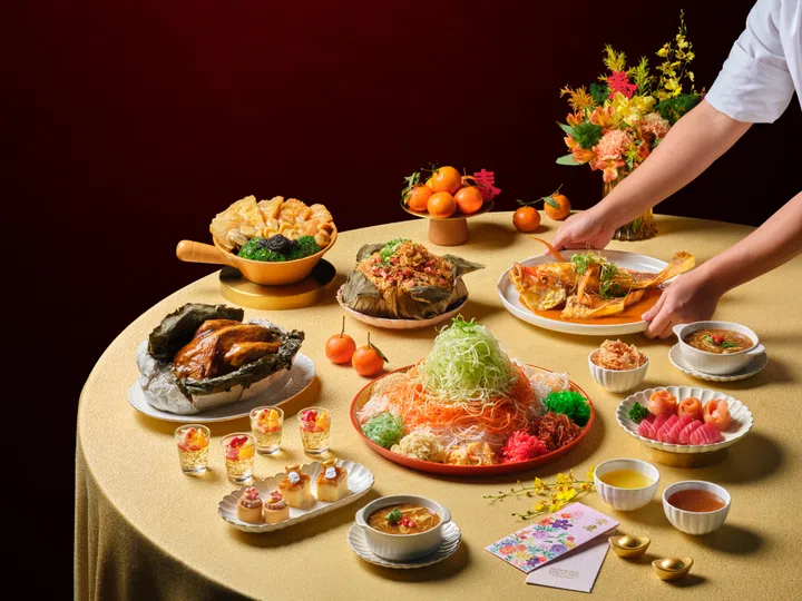 一桌丰盛的新春团圆宴摆满各式中式佳肴，包括色彩缤纷的鱼生拼盘置于中央，搭配烤鸡、整条红烧鱼、海味盆菜、滋补汤品及精致点心等料理。桌上还摆放象征吉祥的柑橘、红包与金色装饰，背景以红色墙面与新春花艺点缀，营造浓厚农历新年喜庆氛围。画面中一双手正端上一盘整条鱼，象征“年年有余”，呈现适合新春期间与家人共享的丰盛团圆盛宴。新加坡大年初一初二照常营业餐厅推荐：8家不打烊好去处（2026马年）