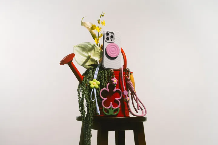 白色涟漪纹理的 iPhone 手机壳，搭配粉色圆形挂件与彩色花朵挂饰，悬挂在红色洒水壶上，周围点缀绿色植物和黄色花朵，整体呈现趣味与花园风格的时尚摆拍。CASETiFY Ripple。高颜值手机壳推荐。