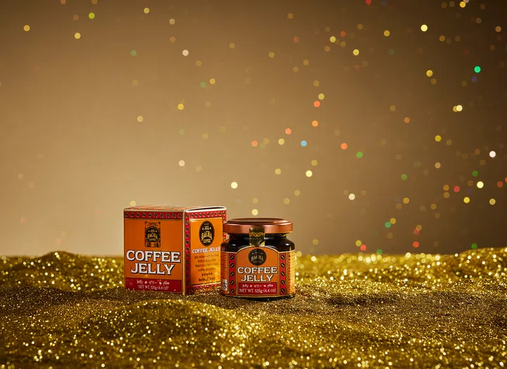 $20以下也能送出好礼：同事交换圣诞礼物精选。画面中摆放着 Bacha Coffee 咖啡冻酱与包装盒，置于金色亮片质地的背景上，后方以节日散景灯点营造温暖圣诞氛围，适用于介绍平价节庆美食小物与交换礼物推荐。