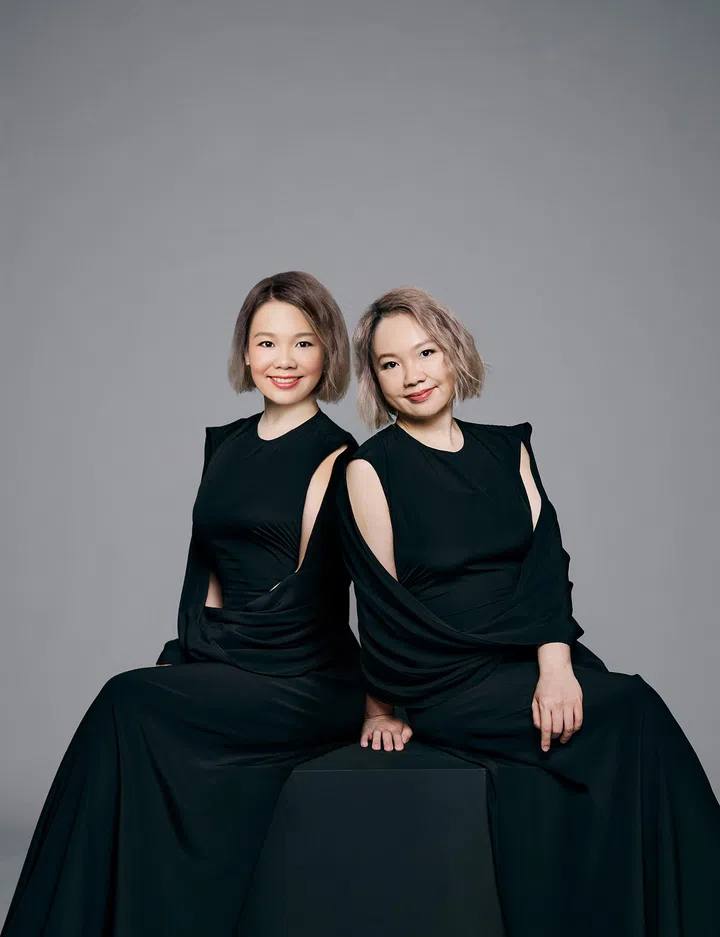 一张棚拍人像照片，两位短发女性并肩坐在深色方形座台上，背景为简洁的浅灰色渐层背景。两人皆身穿剪裁流畅的黑色无袖长礼服，布料在肩部与腰间形成层次感的垂坠线条，整体造型优雅而现代。左侧人物微微侧身，双手自然放在腿上，脸上带着温和的微笑；右侧人物身体略向前倾，一只手轻放在座台边缘，神情从容自信。两人发型皆为及肩短发，发色偏浅棕灰调，妆容自然清新，整体画面呈现出专业、稳重又具亲和力的形象。灯光柔和均匀，突显服装线条与人物轮廓，营造出简约而高级的摄影质感。AI TWINsabrina princessa wang princessa diary 新加坡未来趋势者