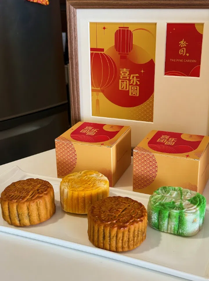 The Pine Garden 2026中秋月饼系列展示图，前景白色长盘摆放四颗不同颜色的月饼，包括两颗金棕色传统烘焙月饼、一颗浅黄色月饼及一颗绿色冰皮月饼。后方陈列橙红色“喜乐团圆”月饼礼盒与品牌卡片，并以木框展示中秋节庆视觉，整体呈现温暖喜庆的送礼氛围。