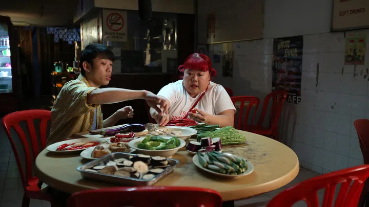 电影《百万红包》中的一幕，两名角色坐在传统咖啡店内的圆桌旁准备料理，桌上摆满各式蔬菜与食材，包括辣椒、茄子、秋葵与苦瓜，红色塑料椅围绕餐桌，整体环境带有老式小贩中心或咖啡店的怀旧氛围，画面呈现出日常生活中的温馨与真实感，呼应电影以平凡日常寻找“开心”的主题。