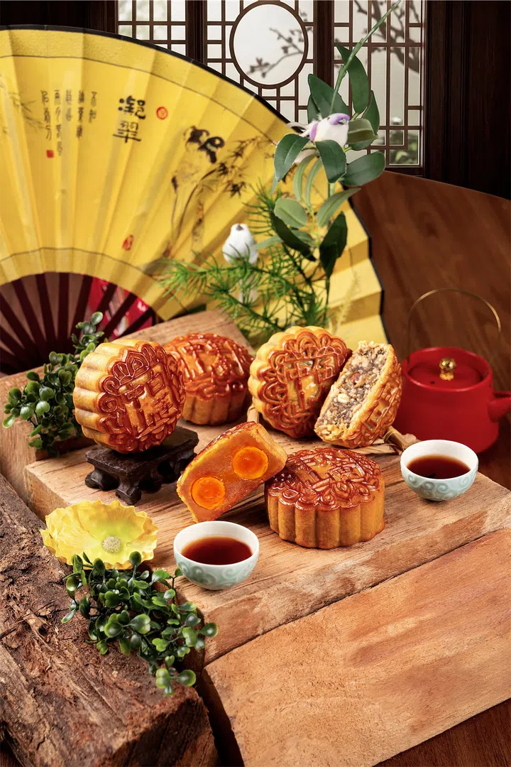 Aloft by Marriott Singapore Novena 2026“中山月饼”系列场景图，以木桌和中式窗格为背景，前景陈列多颗金黄色传统烘焙月饼，其中一颗切开后可见双咸蛋黄内馅，另一颗则展示丰富坚果馅料。画面同时搭配红色茶壶、茶杯、绿植及大型黄色折扇，整体营造带有传统东方元素与节庆气氛的中秋茶席场景。