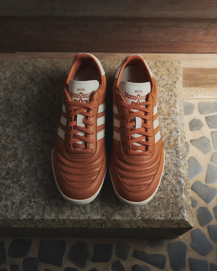 adidas、德国足球俱乐部FC Bayern München与奢侈品牌MCM推出三方联名系列，图片以俯视角度展示一双棕色Mundial运动鞋，整齐摆放在粗糙的灰色石台上。鞋身采用棕色皮革制作，鞋头饰有多层弧形车线，两侧保留adidas经典的奶油白色三条纹，搭配同色系鞋带及浅色鞋底。鞋舌上的奶油白色皮革标签印有MCM品牌标志及月桂叶图案，将adidas的经典运动鞋设计与MCM的奢华品牌元素结合，呈现复古运动与高级时尚交融的风格。