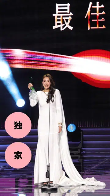 这是一张在《红星大奖》颁奖典礼现场拍摄的照片。舞台中央，刘子绚身穿一袭简洁优雅的白色长礼服，裙摆自然拖地，站在麦克风前，手中举着最佳女主角奖座，脸上带着开心又略显感动的笑容。她的整体造型端庄大方，气质沉稳，成为全场焦点。
舞台背景以黑色为主，上方大字写着“最佳女主角”，搭配红色与白色光影线条，营造出隆重而充满张力的视觉效果。舞台灯光从两侧打下，形成聚光效果，强调得奖者的重要时刻。
画面右侧，一位男艺人身穿黑色西装，正面向刘子绚鼓掌，神情带着欣赏与祝贺。舞台地面呈现镜面反射效果，映出灯光与人物轮廓，让整体画面更具层次感与仪式感。
画面左侧叠加了两个粉色圆形图形，分别写着“独”“家”字样，显示为媒体内容的视觉设计元素。整体画面捕捉了刘子绚获奖当下的荣耀瞬间，气氛温暖而充满掌声与肯定。刘子绚首夺视后：22年7次提名终圆梦，她把时间走成答案