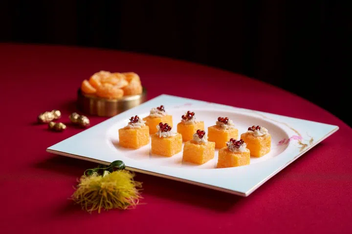 精致摆盘的传统炸蟹肉龙虾浓汤方块整齐排列在白色方形瓷盘上，每块呈金黄色立方体状，外层酥脆，顶部点缀细致蟹肉与晶莹红色鱼子，展现细腻层次与高级质感。瓷盘置于红色桌布上，背景搭配节庆装饰与应景元素，营造浓厚农历新年喜庆氛围，呈现适合新春期间享用的精致中式佳肴。新加坡大年初一初二照常营业餐厅推荐：8家不打烊好去处（2026马年）