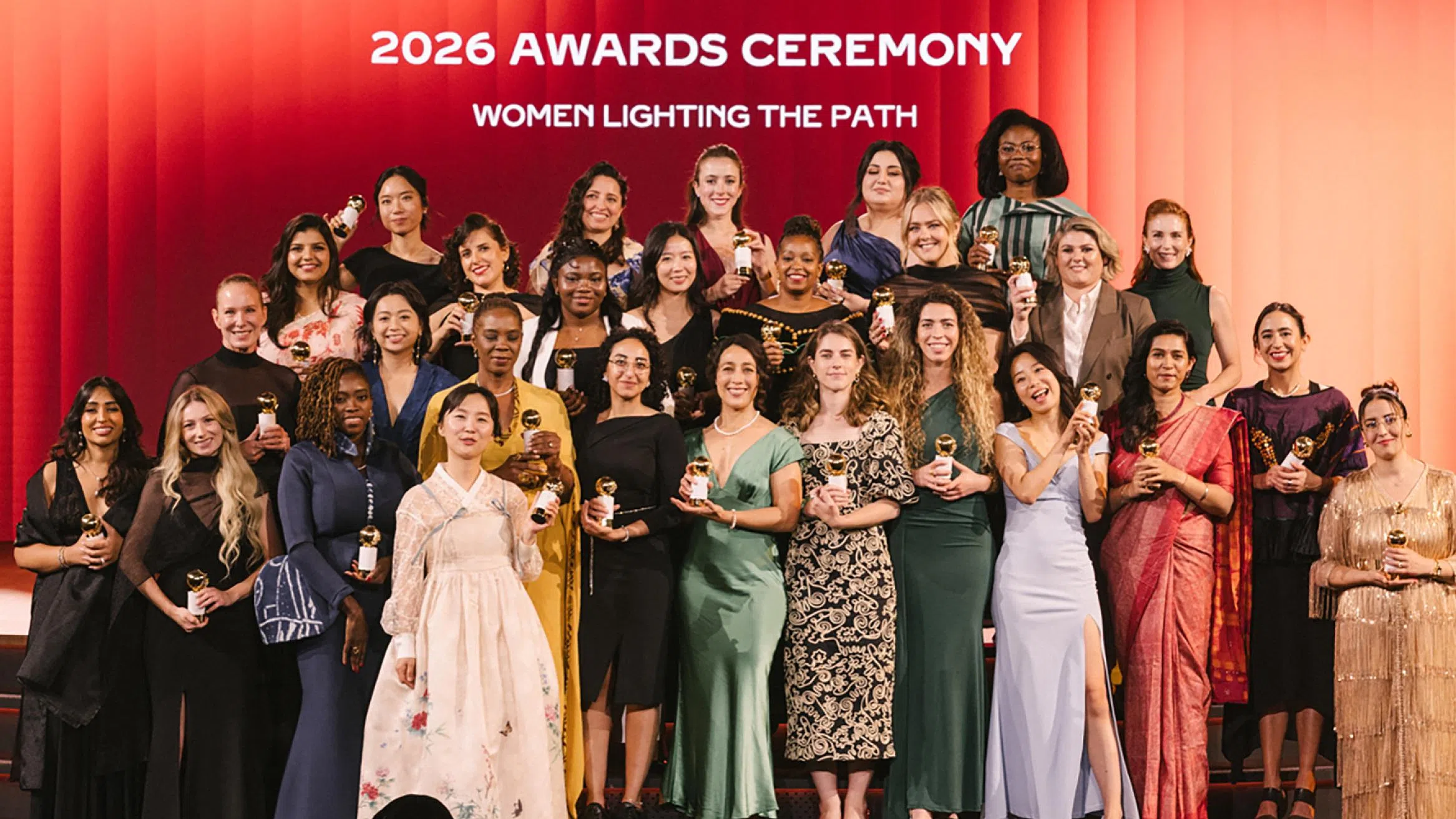 来自19个国家的30位Cartier Women’s Initiative 2026获奖女性创业者齐聚舞台中央，手持象征荣誉的金色奖座合影留念。她们身穿不同国家与文化风格的礼服，站在以红橘色渐层打造的大型舞台前，背景写着“2026 Awards Ceremony”及“Women Lighting the Path”。这场颁奖礼于泰国曼谷朱拉隆功大学举行，同时庆祝Cartier Women’s Initiative成立20周年，现场汇聚来自全球的女性创业者，共同见证这一重要时刻。