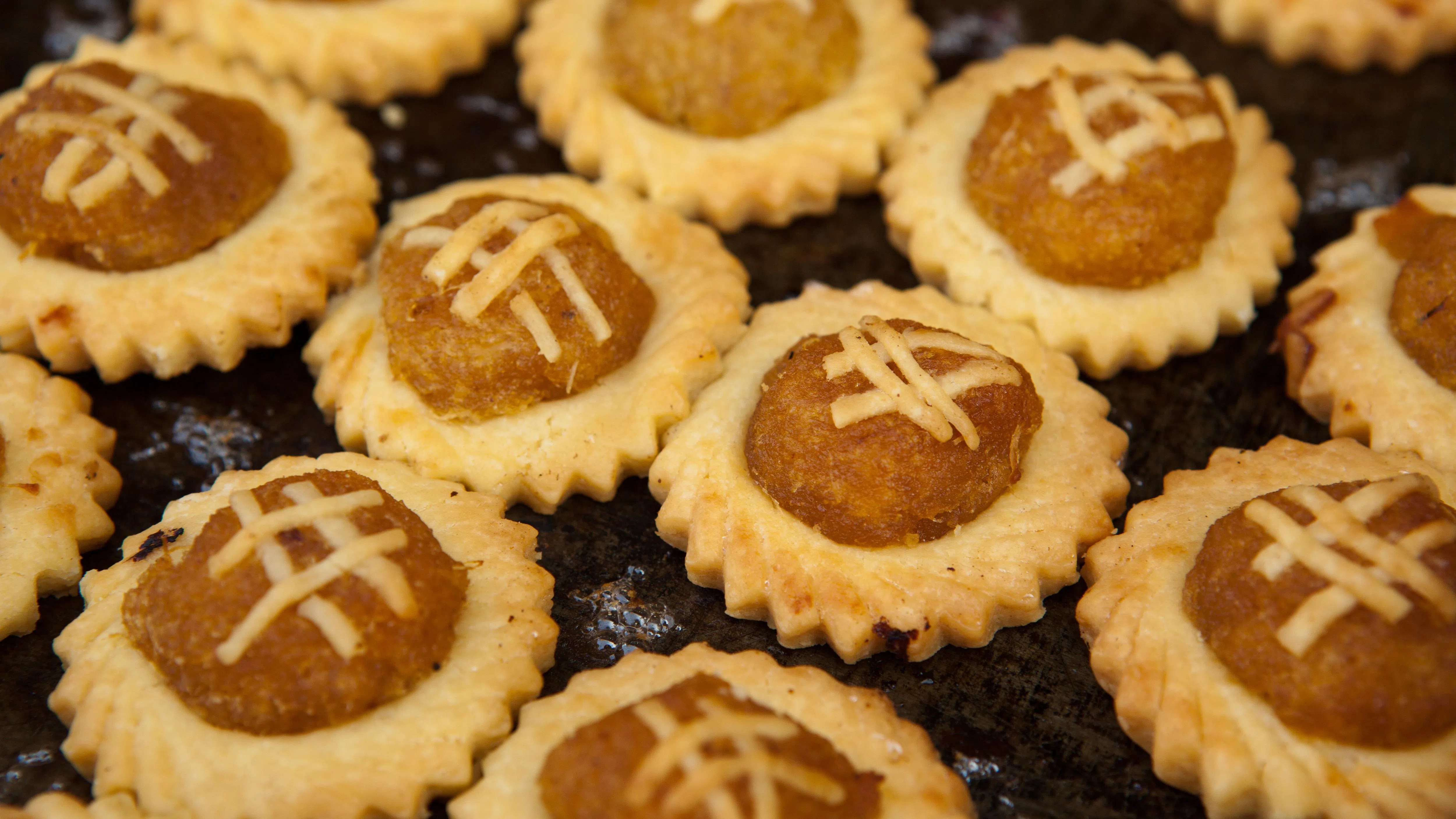 近距离拍摄的一盘凤梨塔，整齐排列在烘焙盘上，每一颗凤梨塔都呈金黄色，带有酥脆的花边饼底，中间放着圆润饱满的凤梨馅，表面饰有细致的交叉面条纹装饰。凤梨馅色泽金黄偏橙，看起来湿润且富有光泽，与浅金色的酥皮形成鲜明对比。整体呈现刚出炉的新鲜状态，质感松脆诱人，是农历新年常见的传统年饼和节庆零食代表之一。新年零食热量排行榜：年饼卡路里大公开，凤梨塔、肉干、鱼皮哪个最高？