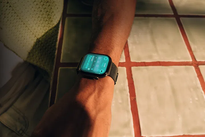 一名用户佩戴Apple Watch Series 12的生活场景图，手腕轻放在白色方砖与红色砖缝组成的桌面上。手表搭配深色金属编织表带，屏幕显示蓝绿色健康资讯界面，并出现“Favorable”状态提示。暖色阳光从侧面照射，突出手腕、表壳与表带的细节，呈现Apple Watch Series 12融入日常佩戴与健康追踪的使用场景。