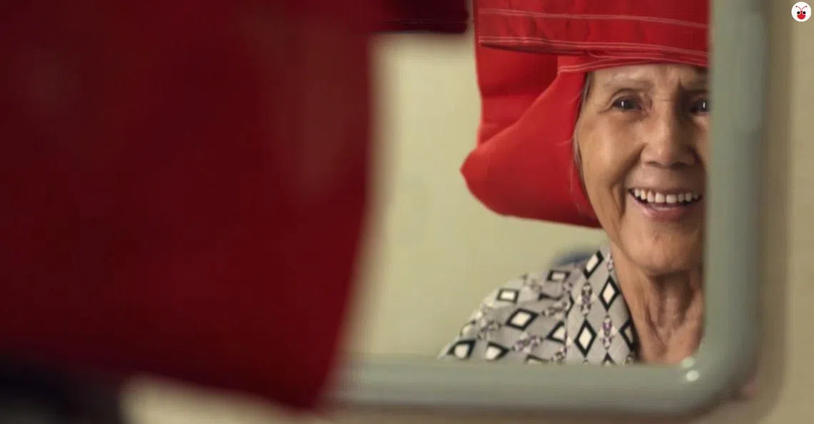 在今年国庆庆典原创影片中，红头巾阿婆胡润心（Woo Yum Sum）看着镜子中戴起红头巾的自己。（影片截图）