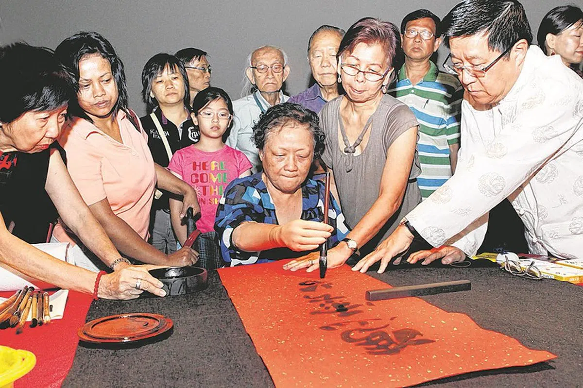  懂得写书法的新加坡人少之又少，而且一般都比较年长。图为本地截断四肢的体障女画家李淑芝（中）2012年在“南艺挥春雅聚和书画展卖会”上当场挥毫响应活动。右一为南洋艺术学院院长朱添寿。 