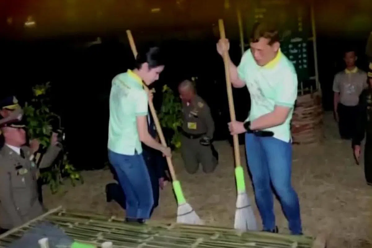  泰王和诗妮娜在镜头前拿起扫把扫地，背景可见几位军官正在行跪礼。（泰国公共电视） 