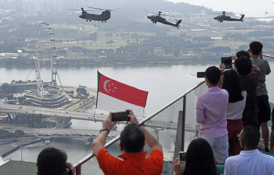8月9日国庆庆祝活动期间，三架直努克直升机悬挂着国旗从滨海湾上空飞过，民众争相拍照。（海峡时报）