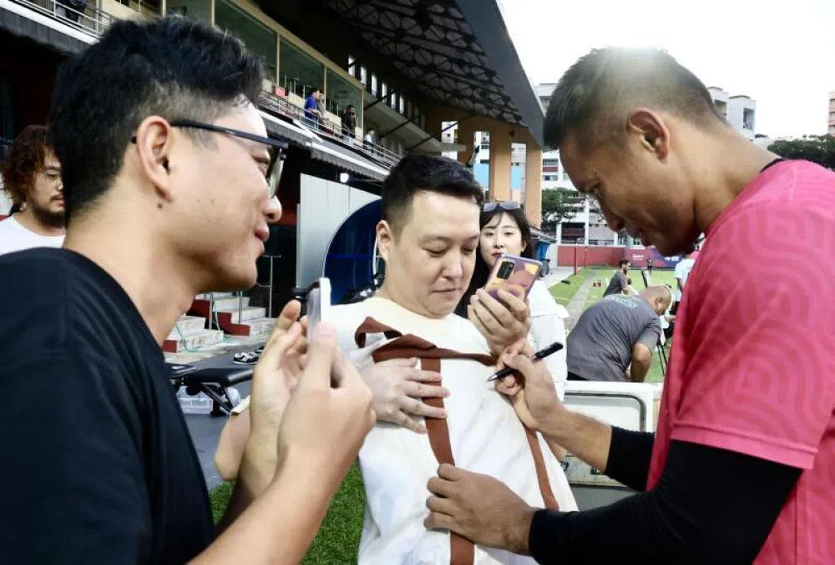 新加坡“雄狮”国家足球队首席门将哈山·桑尼在2024年6月13日在裕廊东体育场参与了球会新潟阿比雷斯的训练，一众中国球迷追到练习场地。图为哈山（红衣O）为特意前来守候的中国球迷签名。（联合早报）