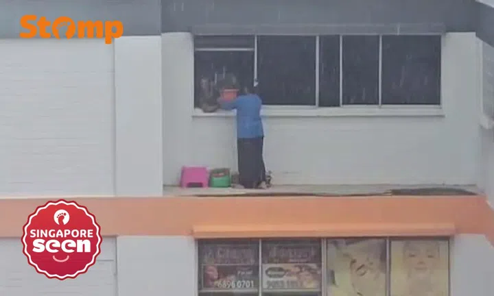 Woman stands on parapet to arrange items outside window during downpour in Bukit Batok