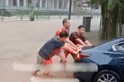 Three men can be seen pushing the car out of the ankle-deep water while drenched in the rain. 