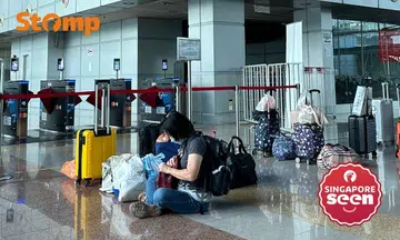 Travellers leave luggage unattended to reserve their spot in the queue, a woman sitting in the centre of the image attends to her own belongings