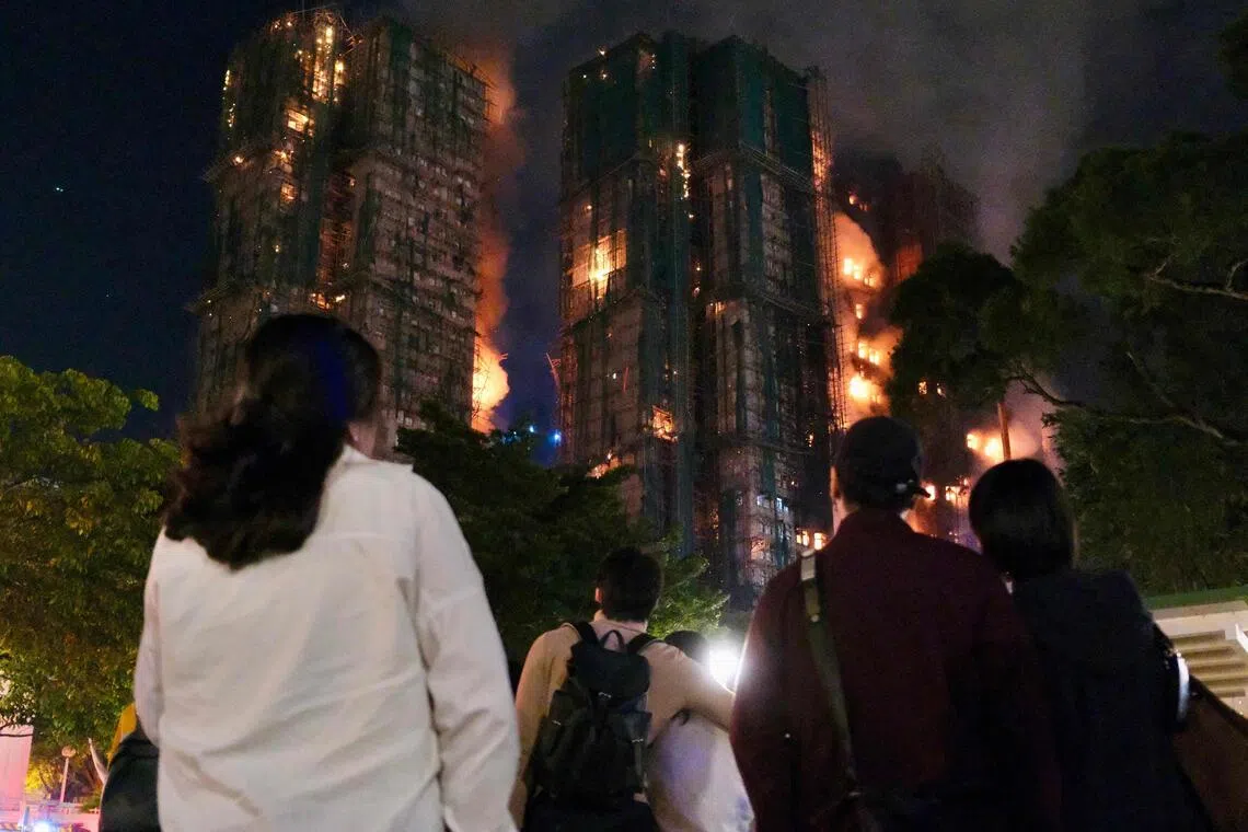 People look on as thick smoke and flames rise during a major fire at the Wang Fuk Court residential estate in Hong Kong's Tai Po district on November 26, 2025. At least 13 people have died as a result of a raging fire that tore through several high-rise blocks in a Hong Kong residential estate on November 26, a government official said. (Photo by Tommy WANG / AFP)
