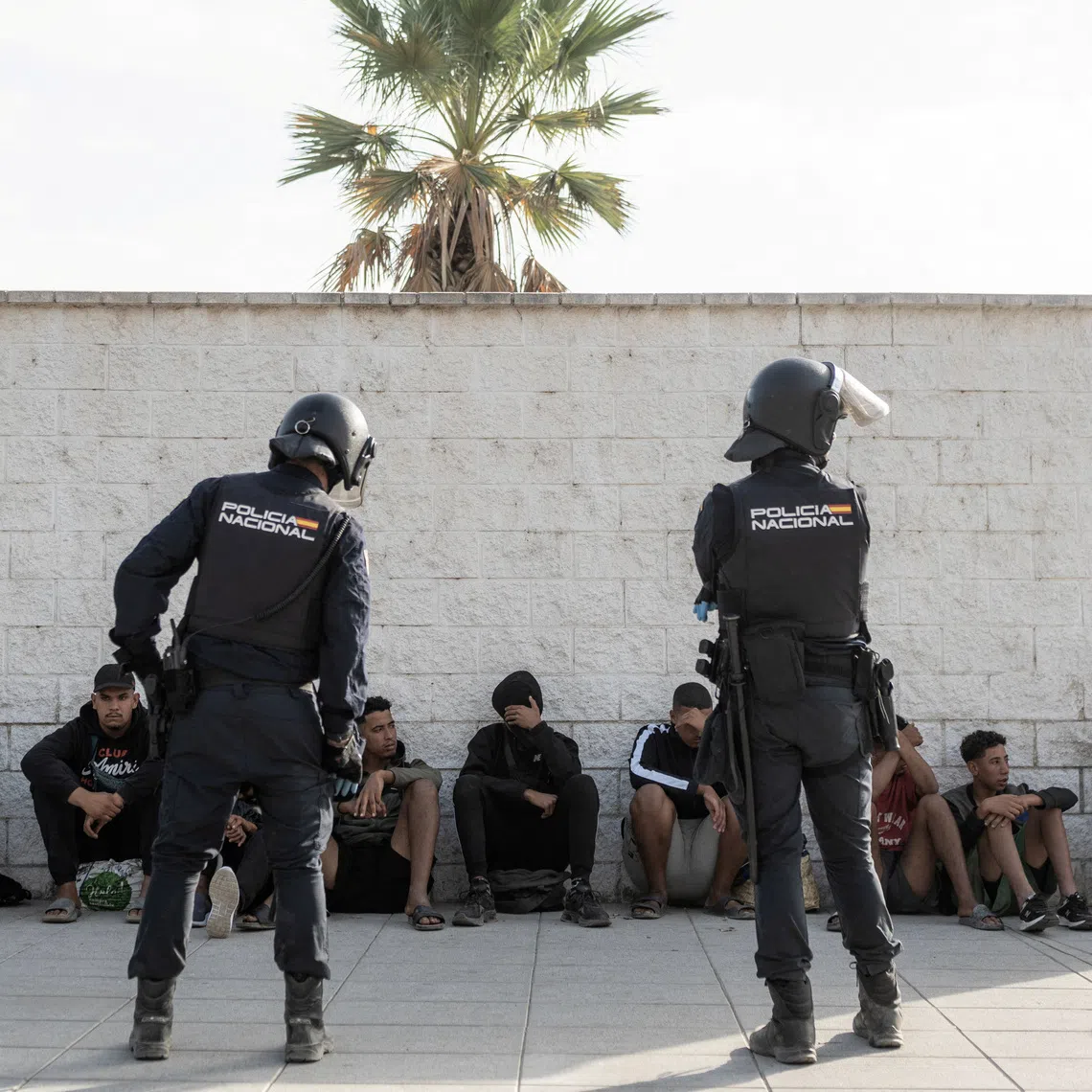 Riot police officers watch a group of migrants before they are transferred to one of the official migrant centers, during an operation to clear part of the makeshift migrant camp at Benitez beach in order to, according to police, establish a security perimeter around Ceuta's desalination plant, following the mass crossing of migrants from Morocco, in Ceuta, Spain, September 27, 2026. REUTERS/Manu Brabo
