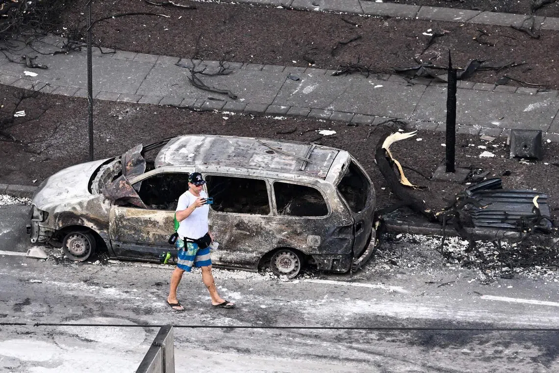 A person walking past a destroyed car in the aftermath of wildfires in Lahaina on Aug 10, 2023. 