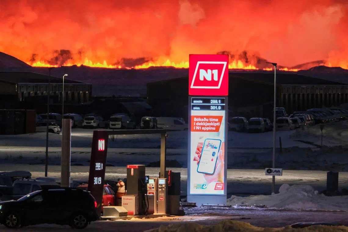 People aree filling up their vehicles at a petrol station as lava and billowing smoke pours out of a fissure during a volcanic eruption near Grindavik, western Iceland on February 8, 2023. A volcanic eruption started on the Reykjanes peninsula in southwestern Iceland on Thursday, the third to hit the area since December, authorities said. (Photo by Kristinn Magnusson / AFP) / Iceland OUT