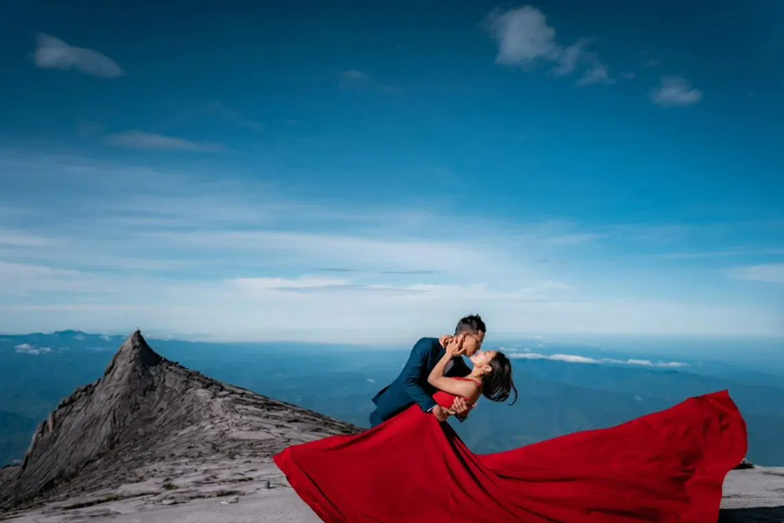 The bride-to-be, dressed in a red sleeveless dress, and her partner, clad in a navy suit with a bowtie, pose with South Peak in the background.
