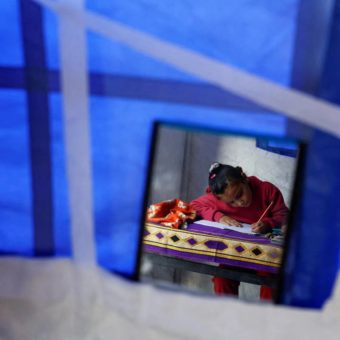 Displaced Palestinian girl Toulin Al-Hindi, 7, is reflected in a hanging mirror as she does her homework inside her family's tent near the Israeli-designated \"yellow line\", in Beit Lahiya, northern Gaza Strip, January 6, 2026. REUTERS/Mahmoud Issa