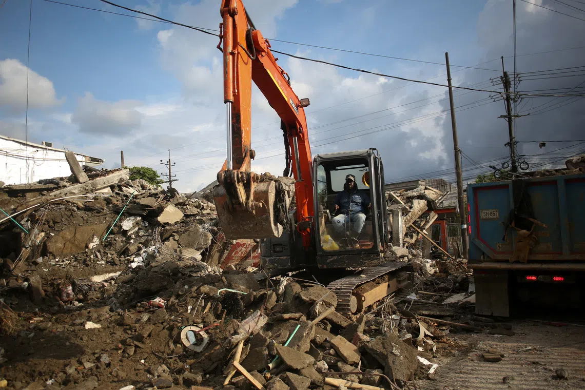 An excavator operator sits in the cab of an orange machine surrounded by piles of construction rubble and debris.