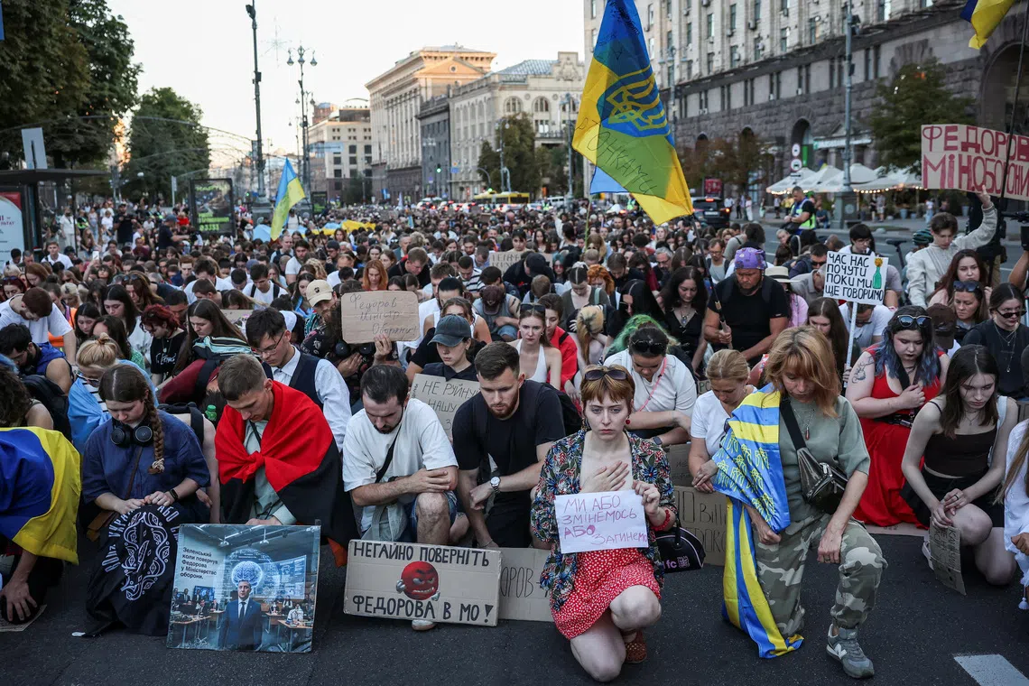 A crowd of protesters kneeling on a street holding signs and blue and yellow flags.