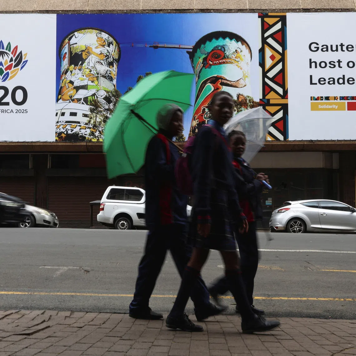 Schoolchildren holding umbrellas walk across the road from a G20 banner as South Africa prepares to host the G20 Summit on November 22-23, in Johannesburg, South Africa, November 18, 2025. REUTERS/Siphiwe Sibeko