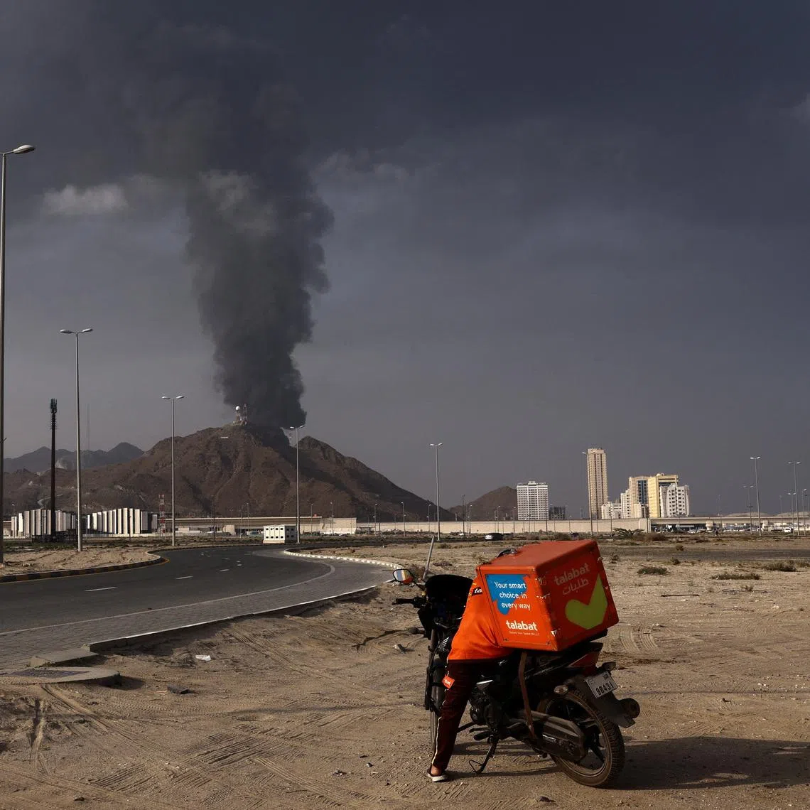 A person stands next to a motorcycle as smoke rises in the Fujairah oil industry zone following a fire caused by debris after interception of a drone by air defenses, according to the Fujairah media office, amid the U.S.-Israel conflict with Iran, in Fujairah, United Arab Emirates, March 3, 2026. REUTERS/Amr Alfiky