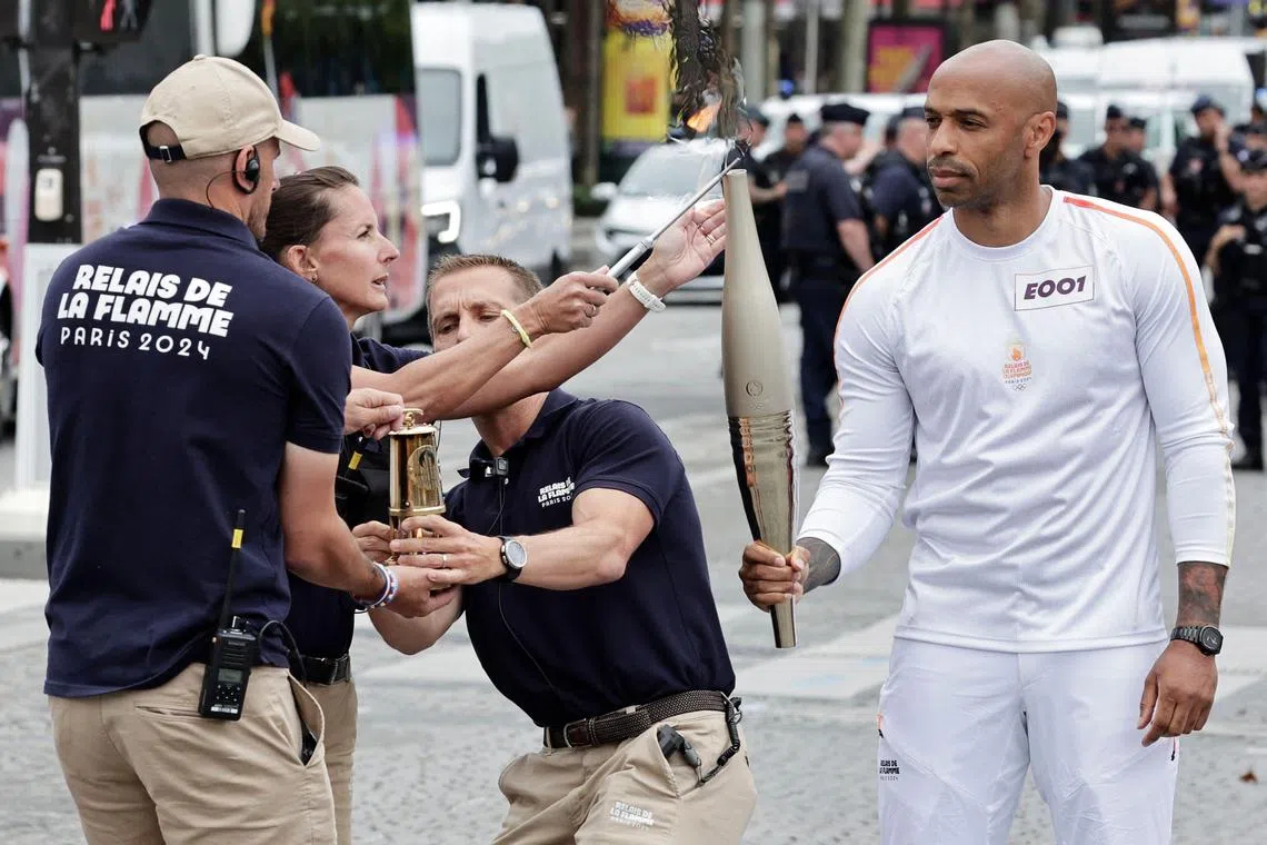 French football Olympics 2024 coach Thierry Henry (R) holds the Olympic torch as officials light the torch during the inauguration of a trunk containing the Olympic torch, custom-designed and manufactured by Louis Vuitton, an LVMH brand partner of the Paris Olympic and Paralympic Games, displayed on the Champs-Elysees avenue, on the sides of the Bastille Day military parade in Paris on July 14, 2024. (Photo by STEPHANE DE SAKUTIN / AFP)