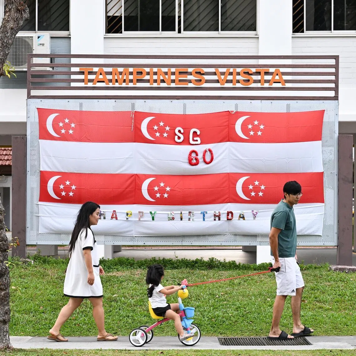Photograph of a young family with a child next to six Singapore Flags put together to wish Singapore Happy Birthday in front of Block 865 Tampines Street 83 on Aug 8, 2025.