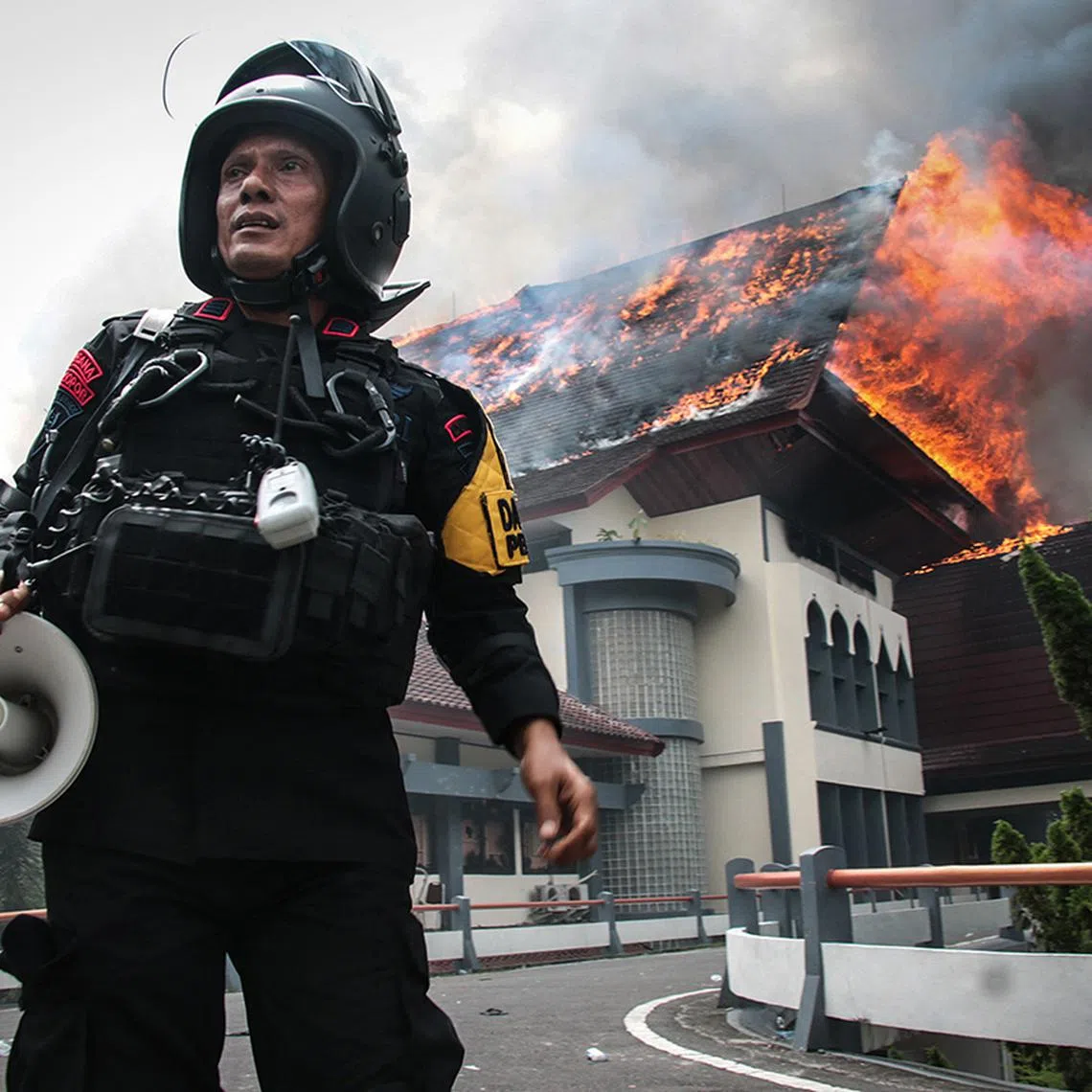 TOPSHOT - A police officer stands with a megaphone as the building of the West Nusa Tenggara Provincial Council office burns after being set on fire by protesters in Mataram on Lombok Island, West Nusa Tenggara, on August 30, 2025. Indonesia was rocked by protests in major cities including the capital Jakarta on August 29 after footage spread of a motorcycle taxi driver being run over by a police tactical vehicle in earlier rallies against low wages and financial perks for lawmakers. (Photo by Sadim / AFP)